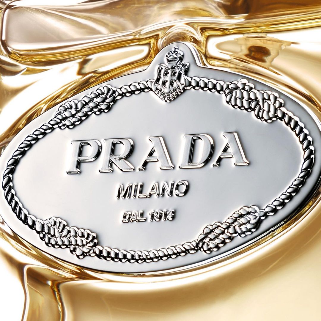 Close-up of a silver Prada logo emblem with rope border on a glossy, reflective gold surface.