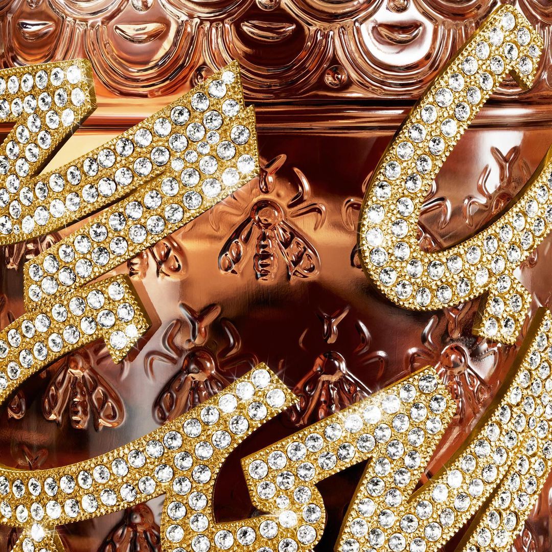 Detailed close-up of an ornate rose gold jar featuring raised honeybee patterns and encrusted rhinestone lettering for a luxurious aesthetic.