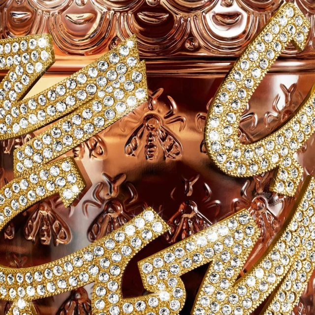 Detailed close-up of an ornate rose gold jar featuring raised honeybee patterns and encrusted rhinestone lettering for a luxurious aesthetic.