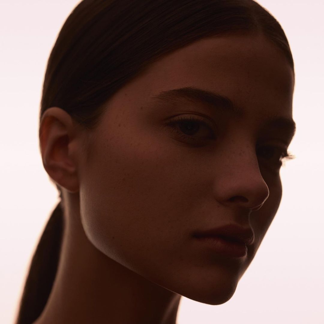 Close-up portrait of an adult female with brown hair pulled back, against a soft pink background. The image has a studio lighting setup.