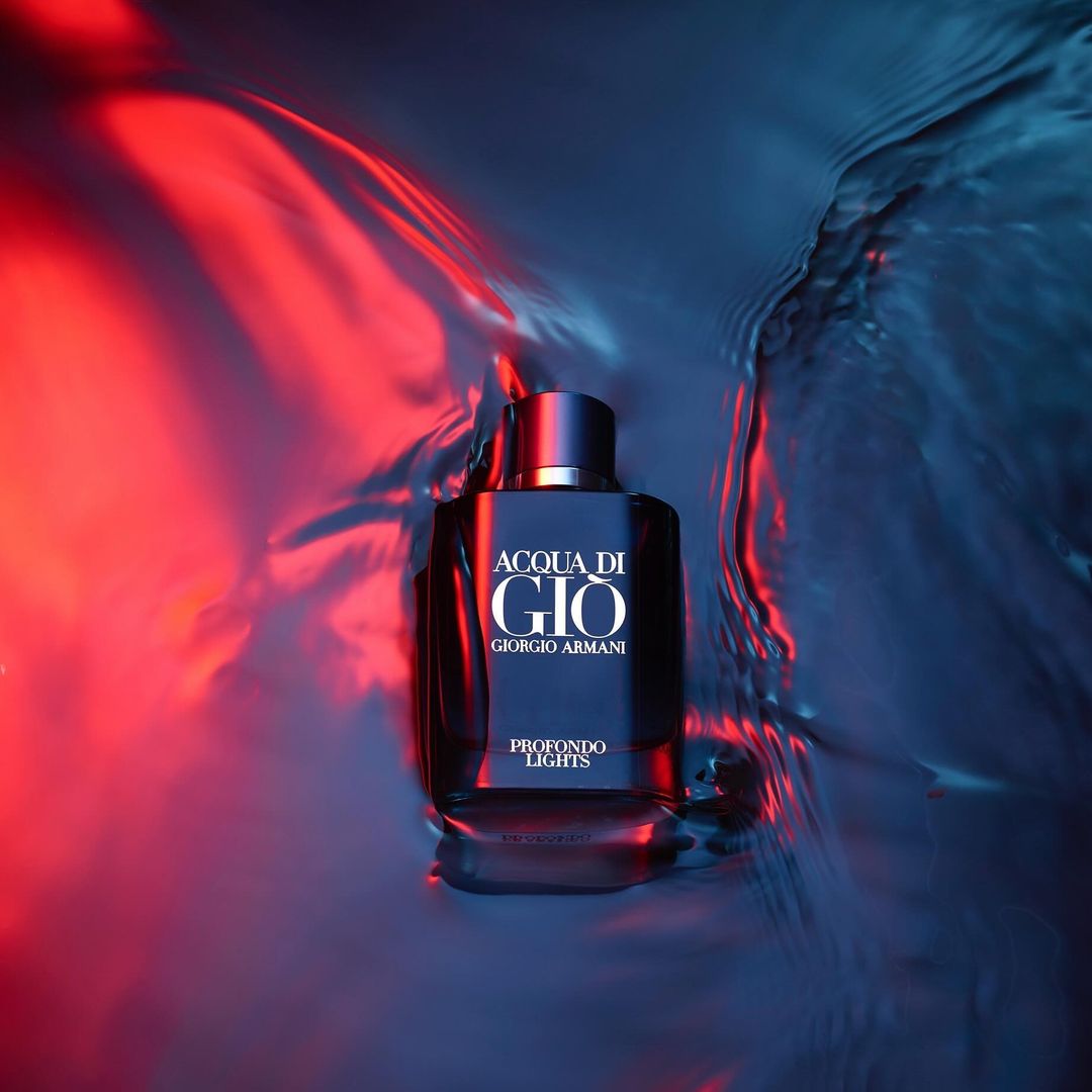 Close-up of a dark perfume bottle titled 'ACQUA DI GIÒ' on a reflective surface with red and blue lighting.