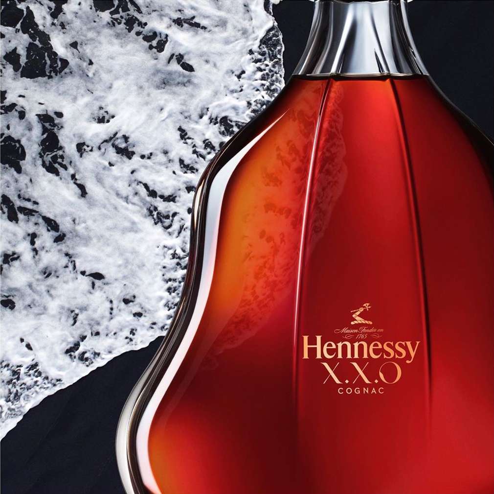 Hennessy X.X.O Cognac bottle close-up against a dark background with contrasting white textured design element.