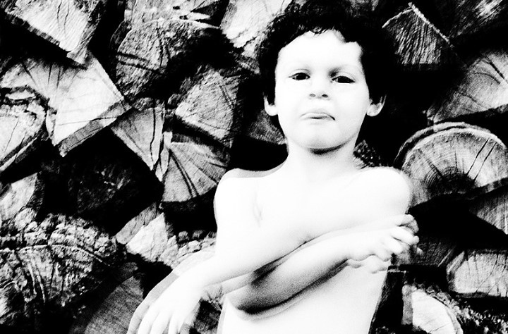 Black and white portrait of a child with curly hair in front of a stack of logs. The child has arms crossed and a neutral expression.