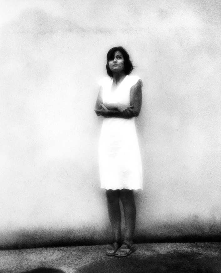 Monochrome full shot of an adult woman in a light dress with arms crossed, standing outdoors against a textured wall.