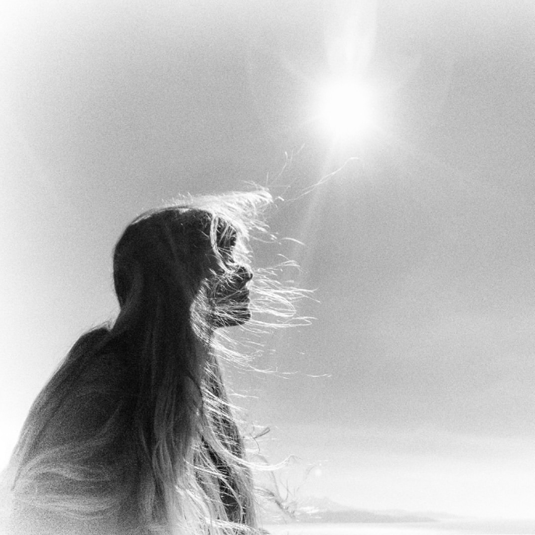 Black and white portrait of a woman's silhouette against a bright sky. Her long hair flows in the wind, creating a sense of movement.