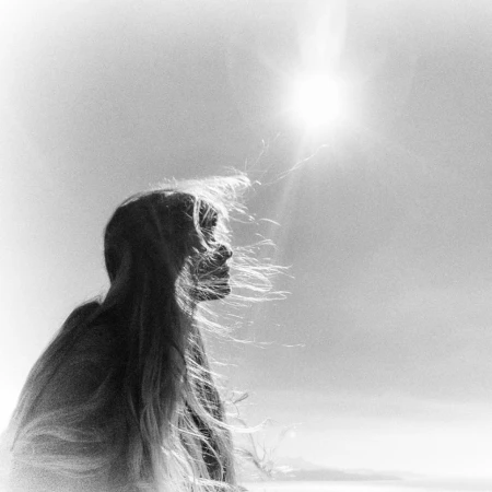 Black and white portrait of a woman's silhouette against a bright sky. Her long hair flows in the wind, creating a sense of movement.