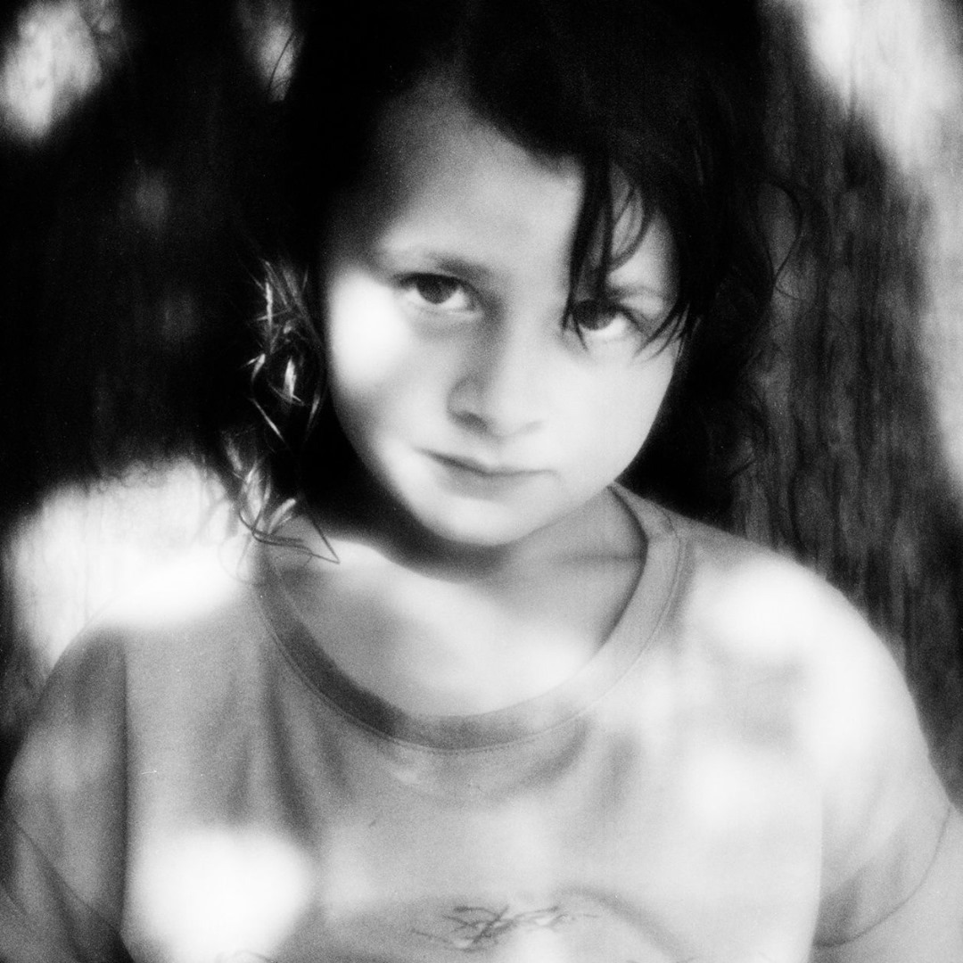Black-and-white portrait of a young girl with dark hair looking at the camera with a tree in the soft-focus background.