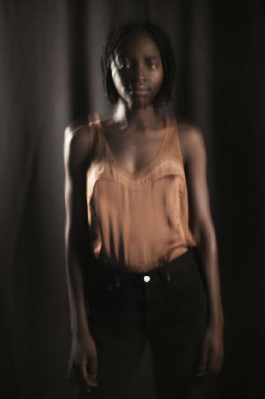 Portrait of a young Black woman in a copper-colored tank top and black pants against a dark curtained background, soft studio lighting.