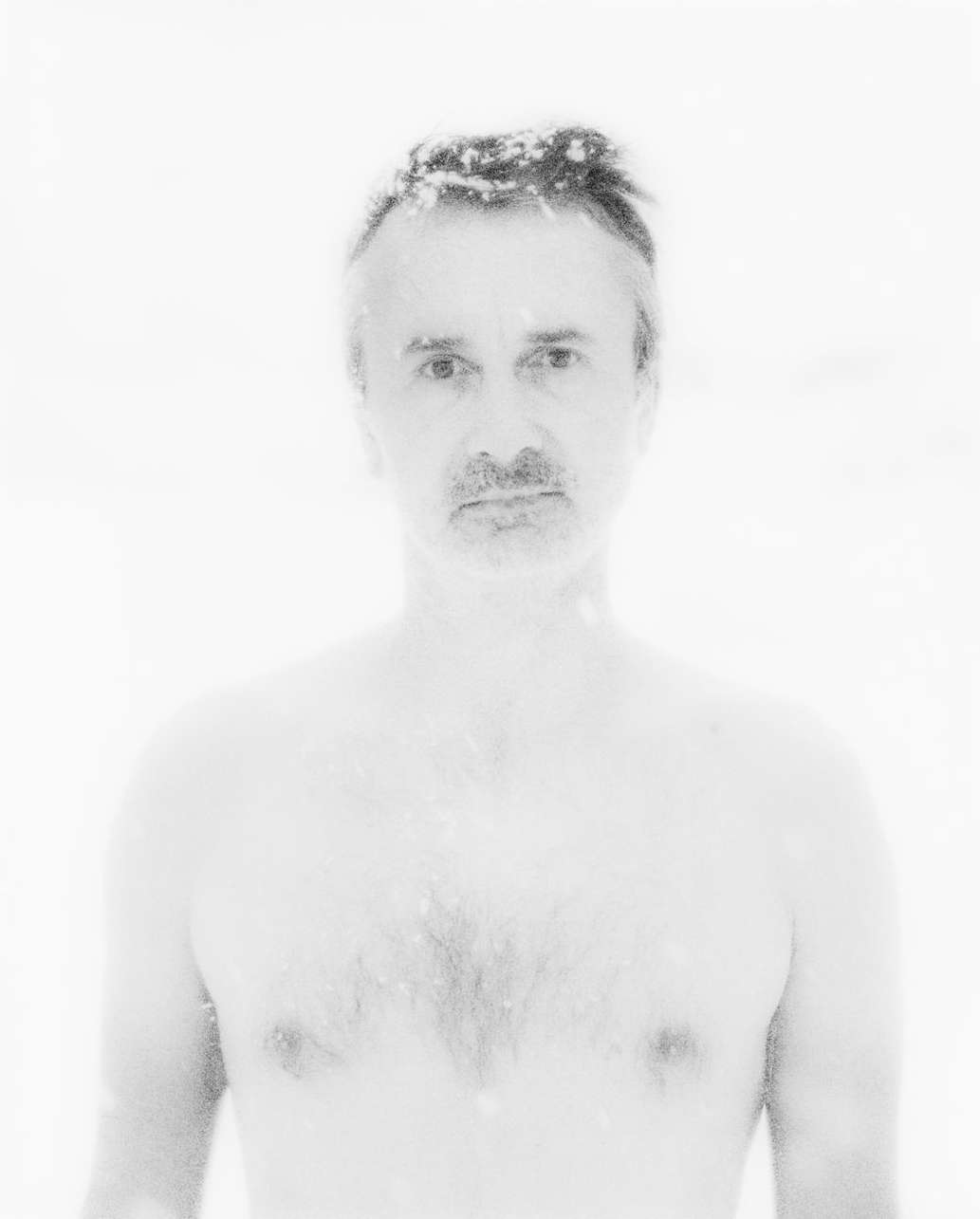 Monochrome portrait of a shirtless adult male with grey hair and stubble, posing against a bright white backdrop, captured in a studio setting.