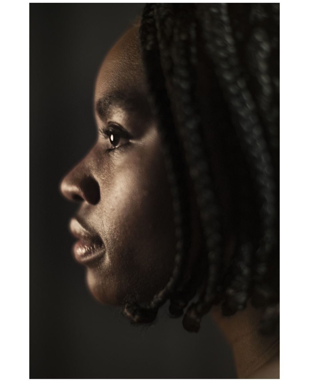 Close-up portrait of a young African-American woman with braided hair in profile, bathed in soft lighting against a dark background.