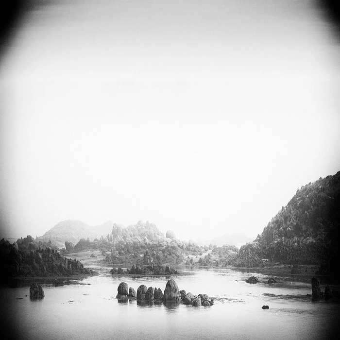 Monochrome landscape photograph of rocky outcrops emerging from tranquil water under a hazy, bright sky.