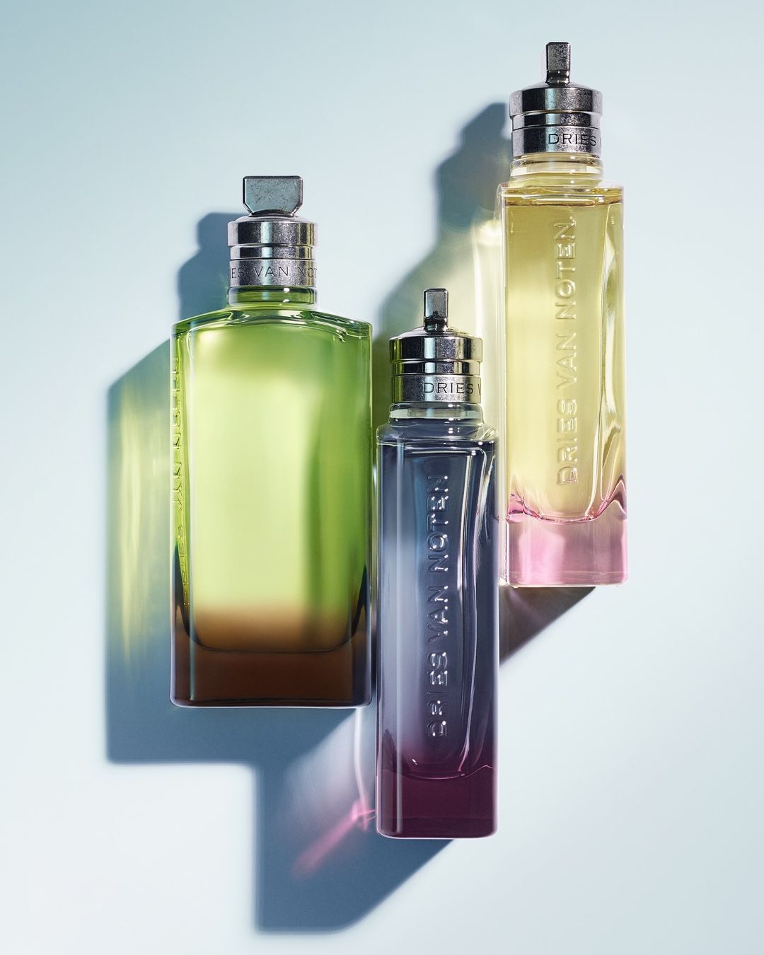 Three Dries Van Noten perfume bottles in varying sizes and gradient colors, arranged against a gradient blue background.