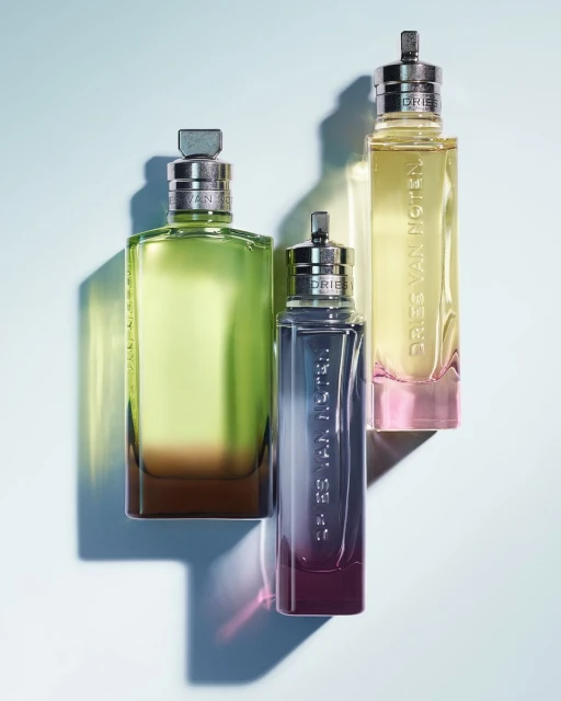 Three Dries Van Noten perfume bottles in varying sizes and gradient colors, arranged against a gradient blue background.