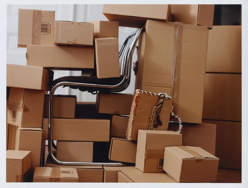 A chrome chair is piled high with cardboard shipping boxes in a bright indoor space, hinting at moving or storage needs.