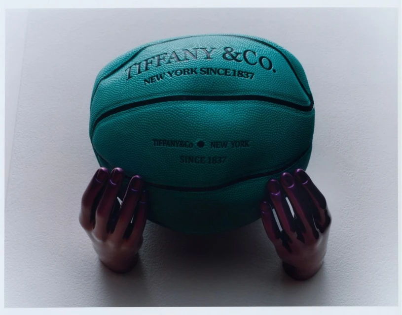Teal-colored basketball with Tiffany & Co. logo, held by two glossy purple mannequin hands against a plain light-colored background.