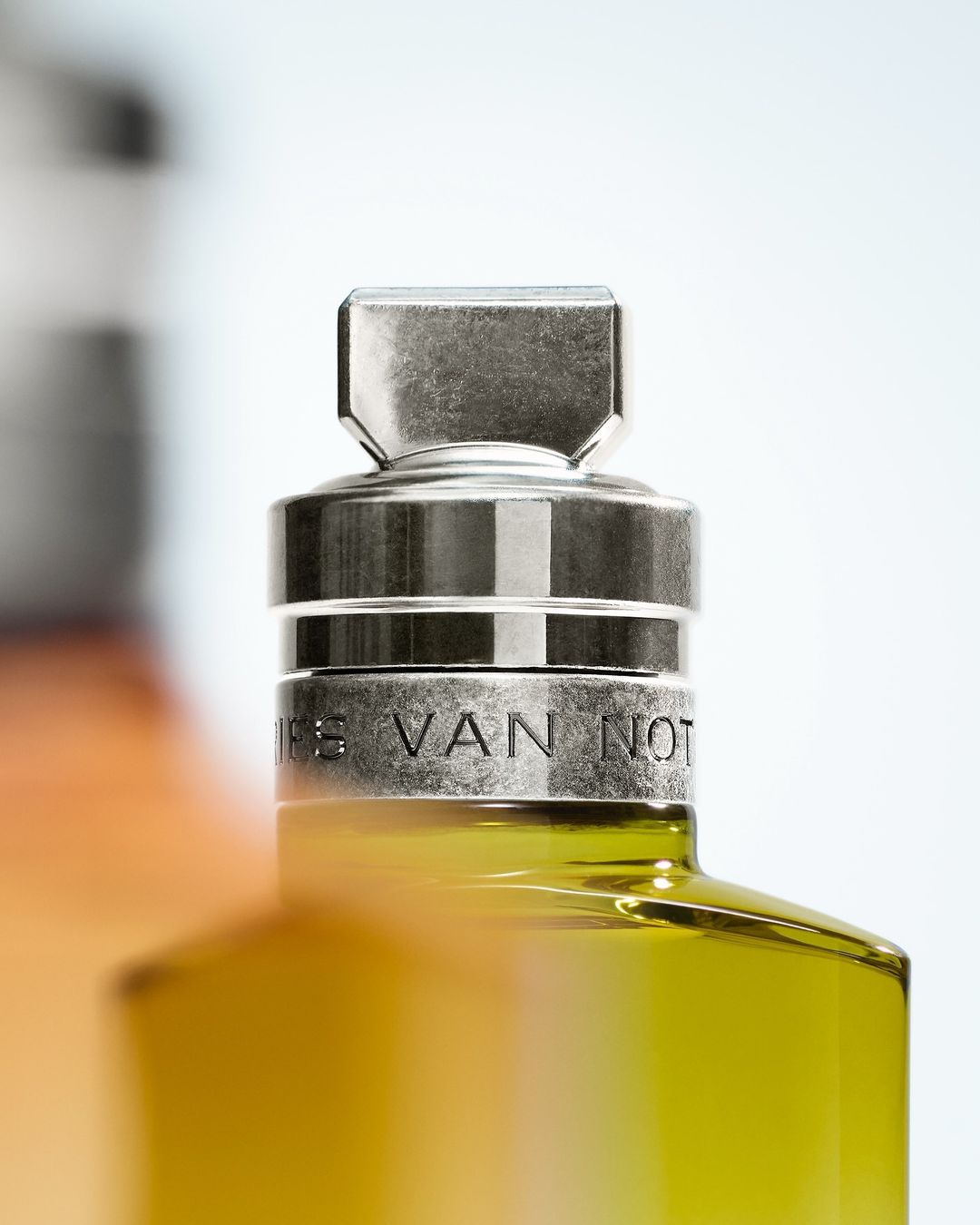 Close-up of a partially visible perfume bottle with a metal cap and 'DRIES VAN NOTEN' engraved on it, against a bright background.