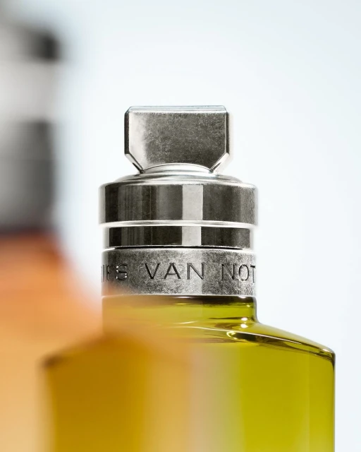 Close-up of a partially visible perfume bottle with a metal cap and 'DRIES VAN NOTEN' engraved on it, against a bright background.