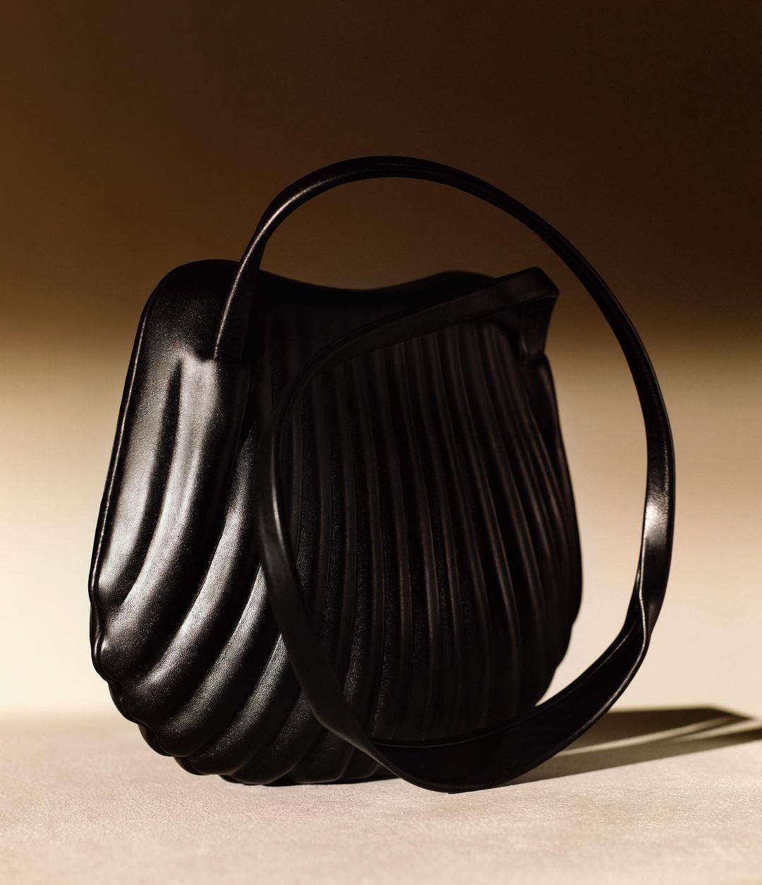 Elegant black pleated handbag with a unique looped handle, presented on a light surface against a softly lit, graduated brown background.