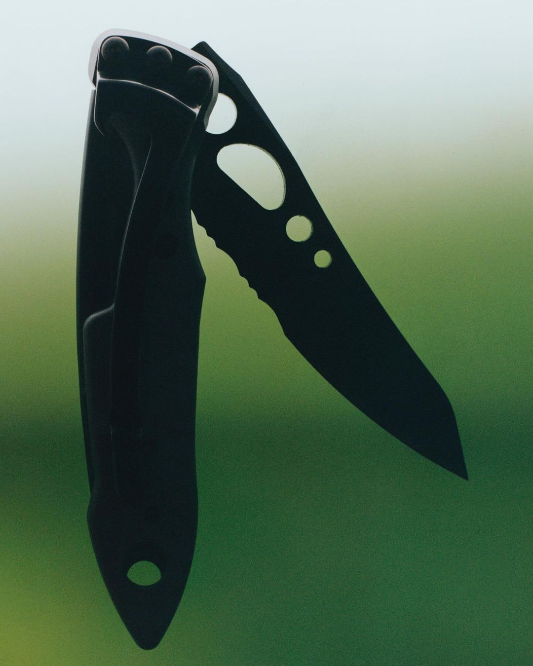 Close-up of a black folding knife with a serrated blade, showcasing its design and construction against a green background.