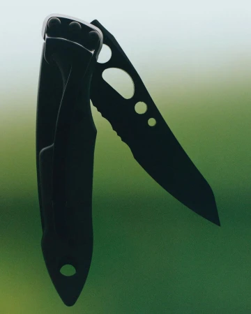 Close-up of a black folding knife with a serrated blade, showcasing its design and construction against a green background.