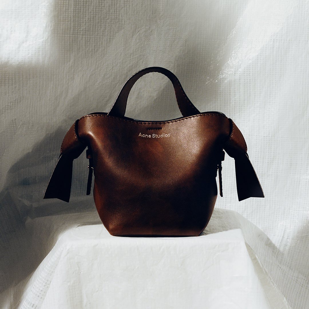 A brown leather Acne Studios handbag sits on a white tablecloth against a white backdrop, emphasizing a minimalist studio aesthetic.