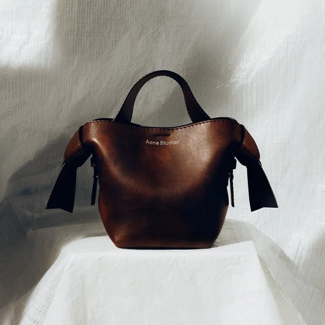 A brown leather Acne Studios handbag sits on a white tablecloth against a white backdrop, emphasizing a minimalist studio aesthetic.
