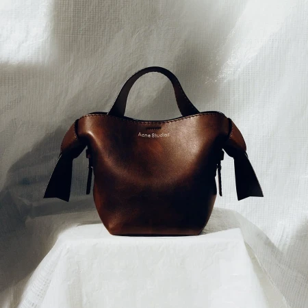 A brown leather Acne Studios handbag sits on a white tablecloth against a white backdrop, emphasizing a minimalist studio aesthetic.