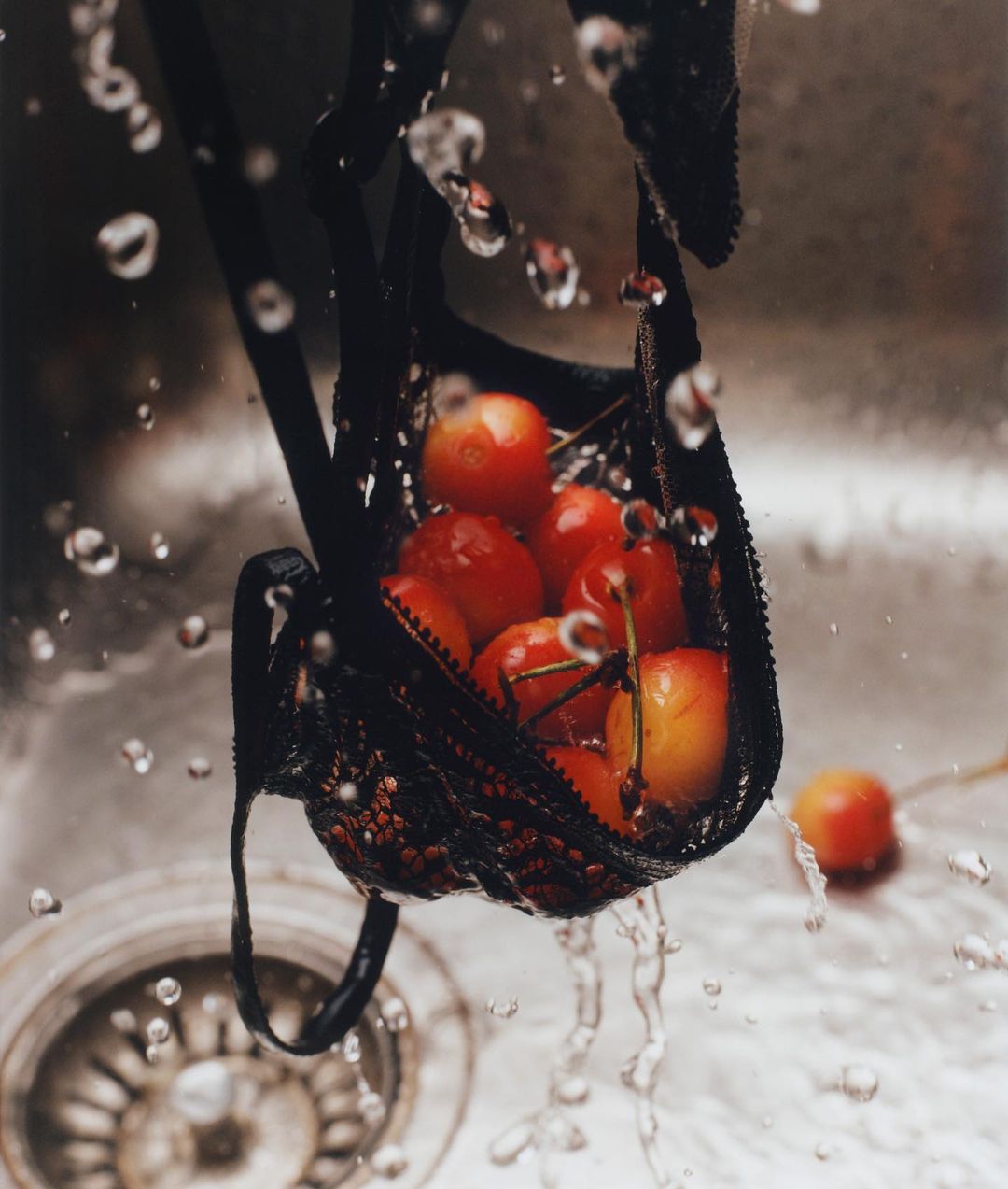 Red cherries filling a black bra being rinsed in a stainless steel sink with water splashing, close-up, overhead shot, and high-key lighting.