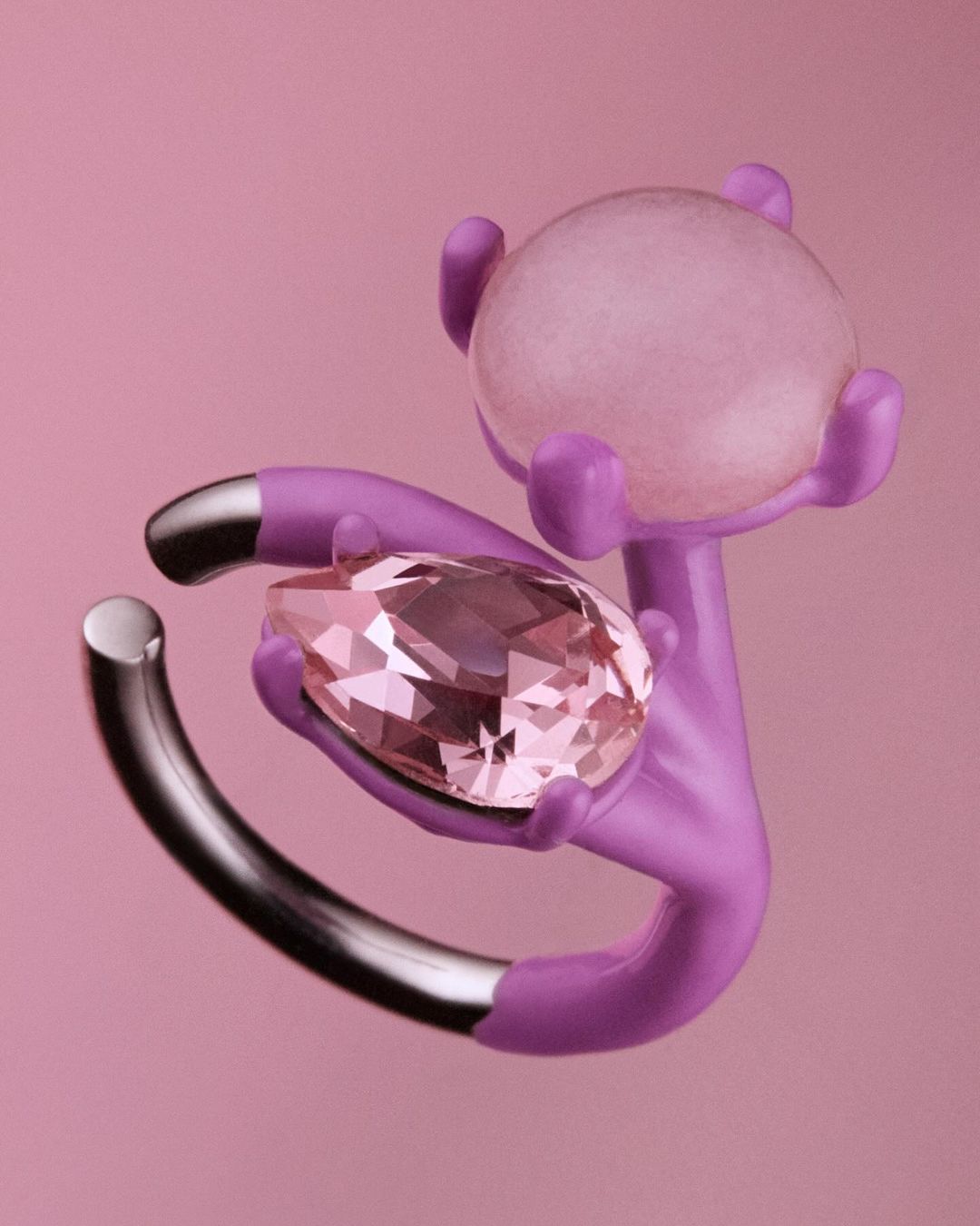 Still life featuring a pear-cut gemstone, a lavender abstract hand sculpture, and a frosted spherical object on a pink backdrop.