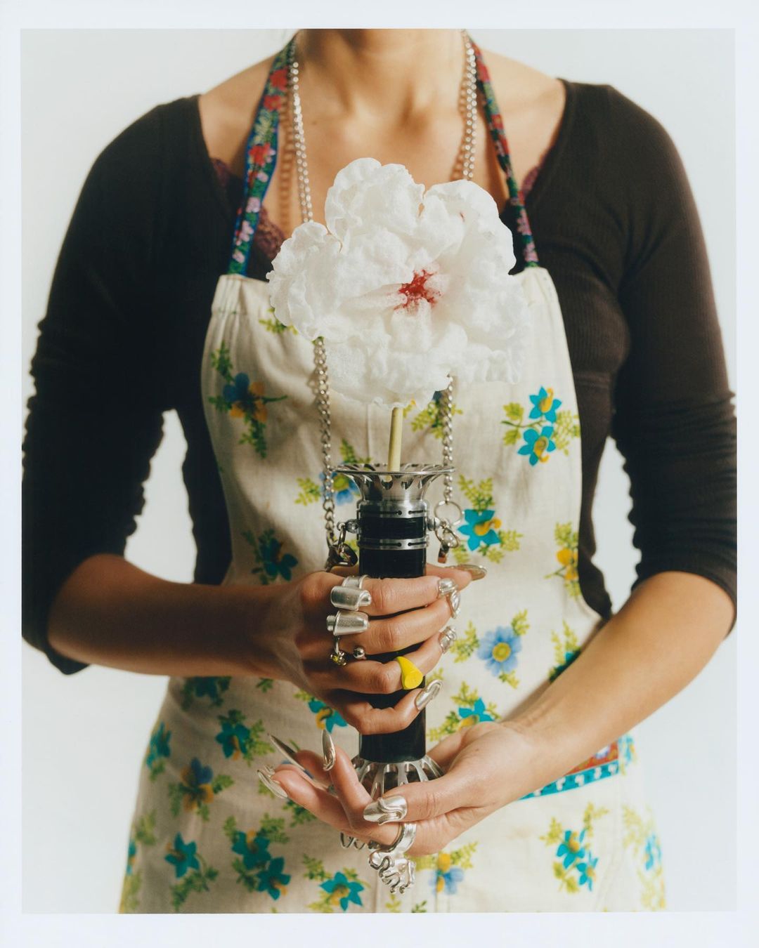 Waist-up shot of a person holding a large flower in a decorative holder while wearing an apron and multiple rings, against a white backdrop.
