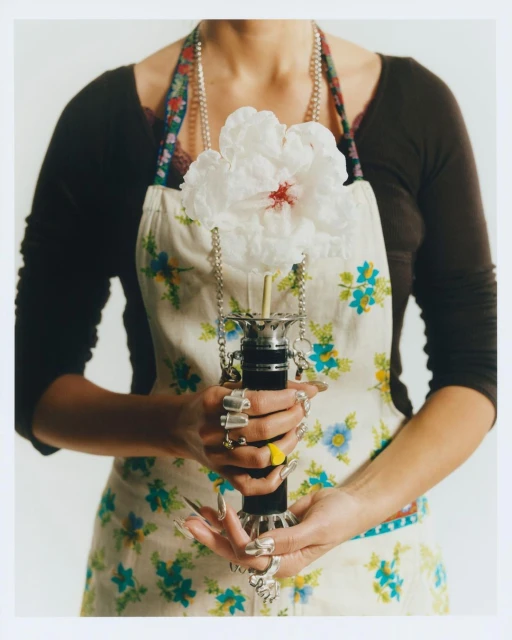 Waist-up shot of a person holding a large flower in a decorative holder while wearing an apron and multiple rings, against a white backdrop.