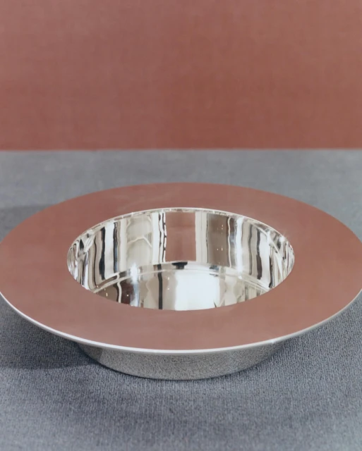 Elevated close-up of a reflective silver bowl with a wide rim, set against a muted backdrop of gray and earthy red tones.