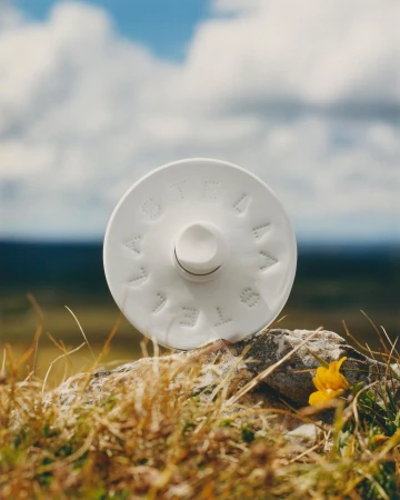 Round white ceramic bottle perched on a rocky ground with tall grass against a dramatic cloudy sky.