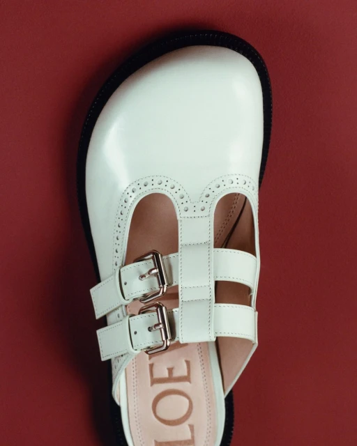 Overhead shot of a white leather mule with double straps and silver buckles, set against a deep red background for fashion.