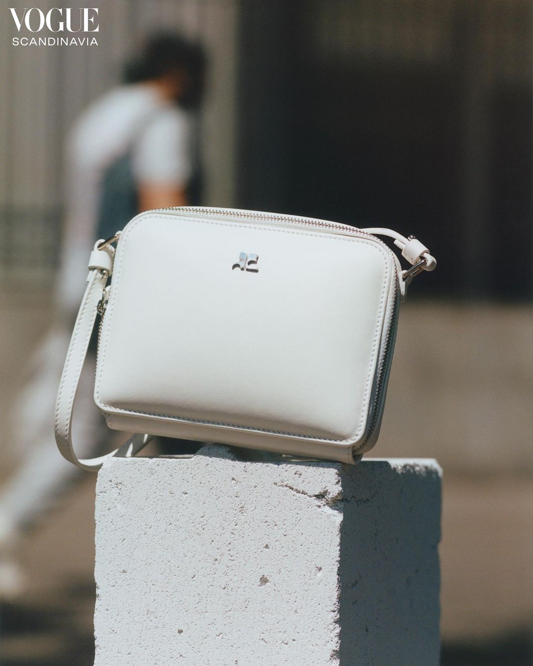 White crossbody bag with silver hardware atop a concrete block outdoors. Person walking in background. 'Vogue Scandinavia' logo visible.