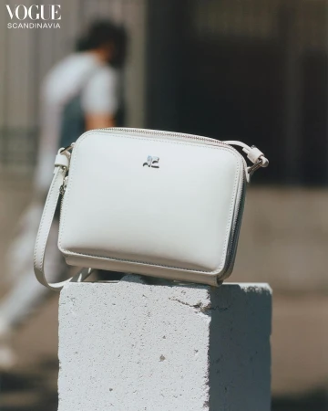 White crossbody bag with silver hardware atop a concrete block outdoors. Person walking in background. 'Vogue Scandinavia' logo visible.