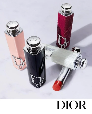 Four Dior lipstick tubes in pink, black, burgundy, and white arranged on a light marble surface with silver hardware details.