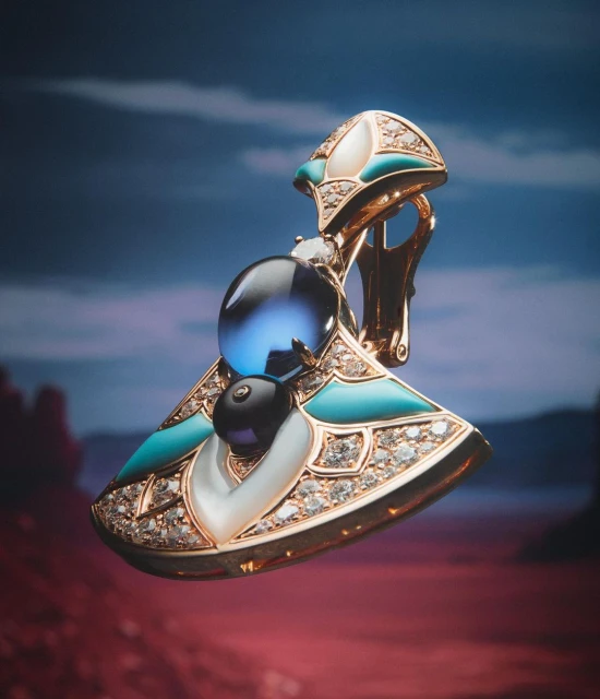 Studio shot of an earring featuring blue and black round gemstones and inlaid diamonds in a gold setting, against a blurry desert backdrop.