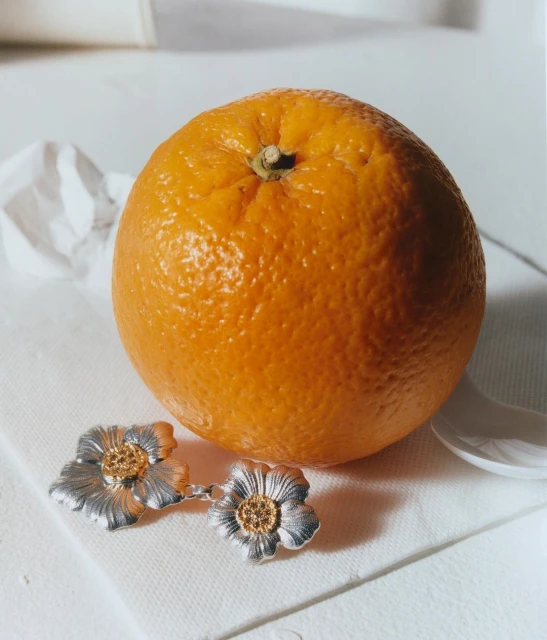Still life close-up of an orange with silver flower earrings displayed on a white surface, creating a bright, minimalist composition.