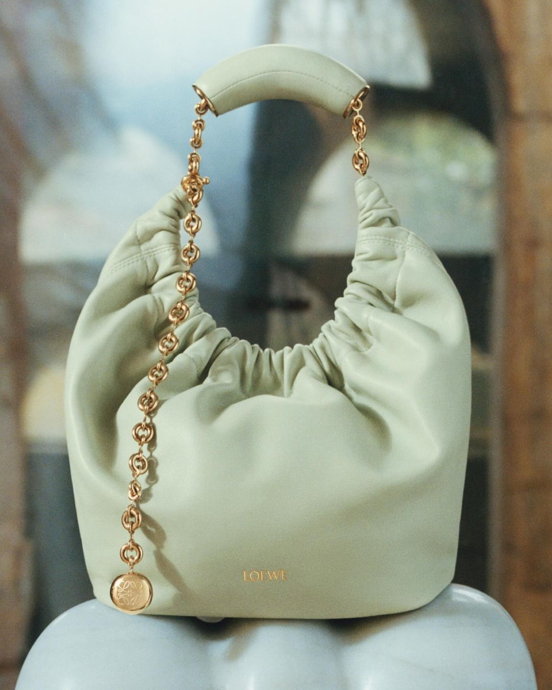 Loewe Luna handbag in light green leather with chain handle and gold emblem on a white marble surface in an indoor setting.