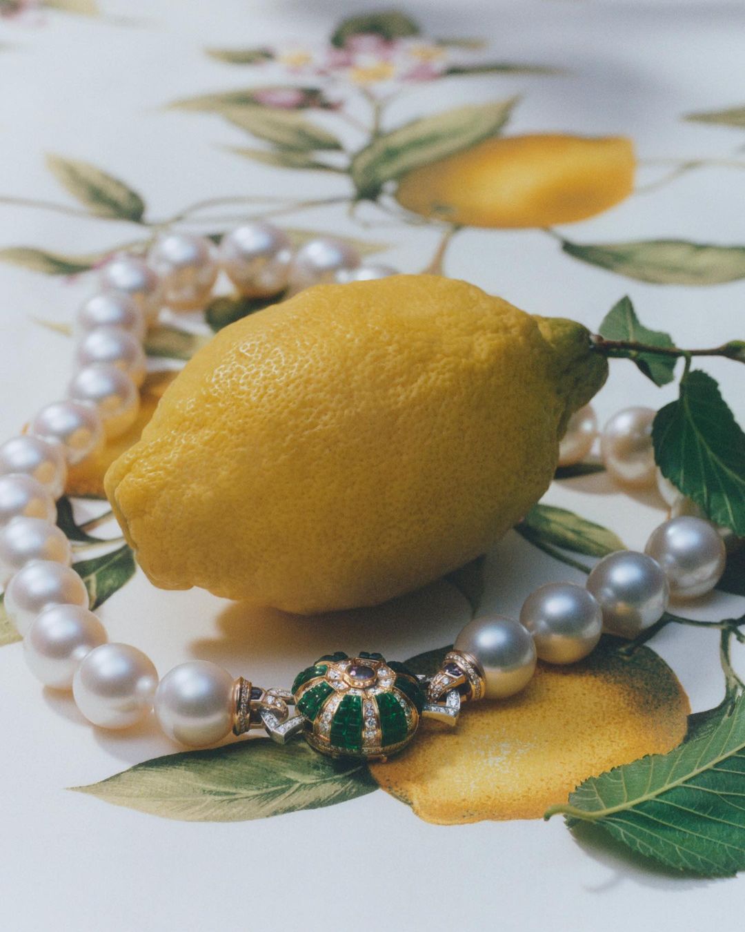 A pearl necklace with an emerald clasp resting beside a whole yellow lemon on a patterned botanical tabletop.