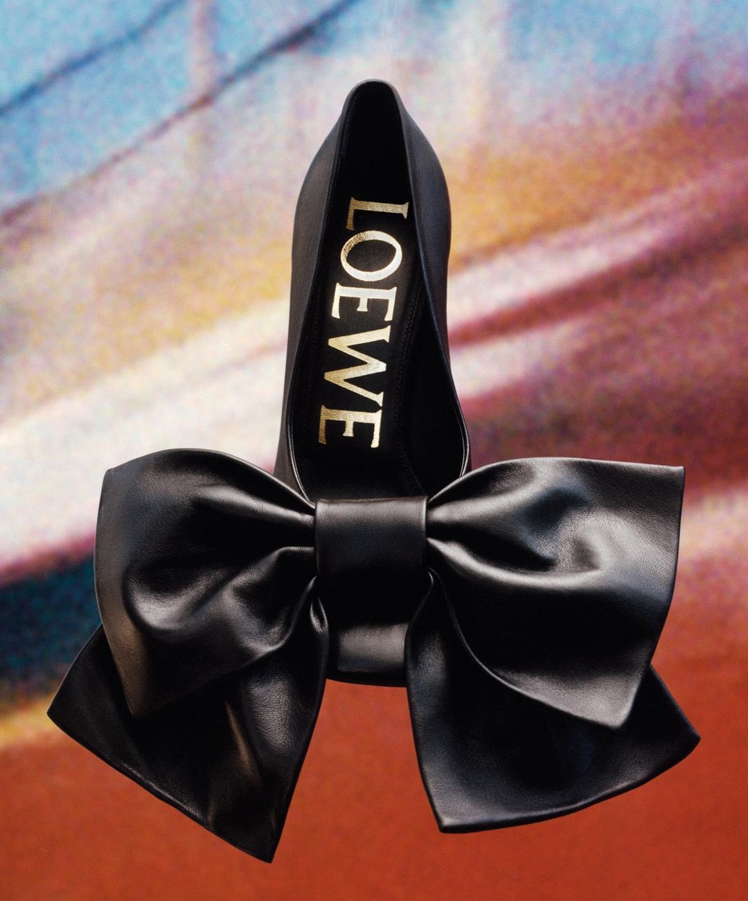 Elevated view of a Loewe black leather pump with an oversized bow, presented against a textured, painterly backdrop, showcasing luxury footwear.