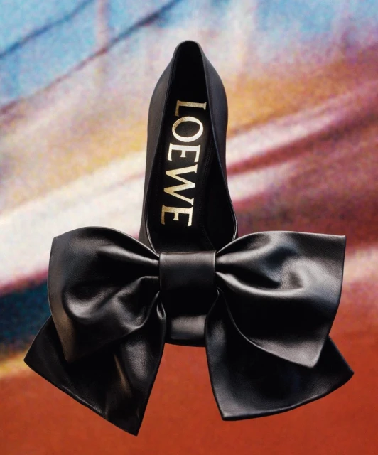 Elevated view of a Loewe black leather pump with an oversized bow, presented against a textured, painterly backdrop, showcasing luxury footwear.