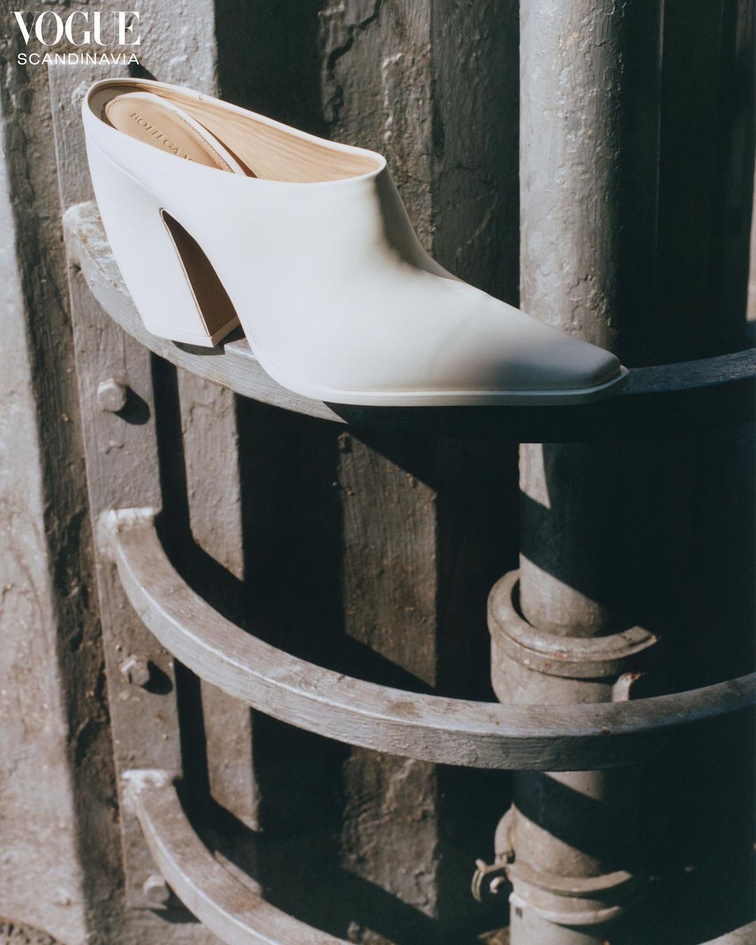 Cream-colored heeled mule is featured on a weathered, grey, multi-tiered column in soft light for a fashion editorial spread.
