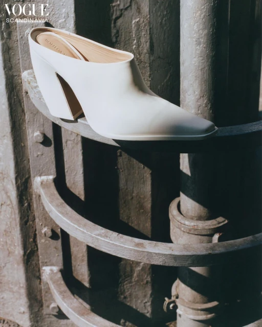 Cream-colored heeled mule is featured on a weathered, grey, multi-tiered column in soft light for a fashion editorial spread.