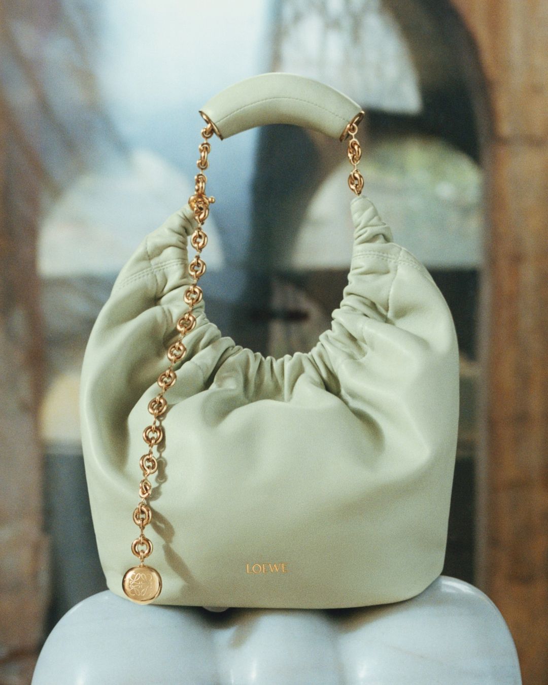 Elegant Loewe purse in light green leather with a gold chain detail, displayed against a neutral architectural backdrop.