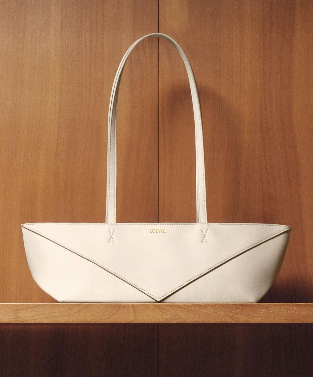 A cream-colored LOEWE envelope bag is presented on a shelf against a wooden wall. The bag features long straps and a unique, folded design.