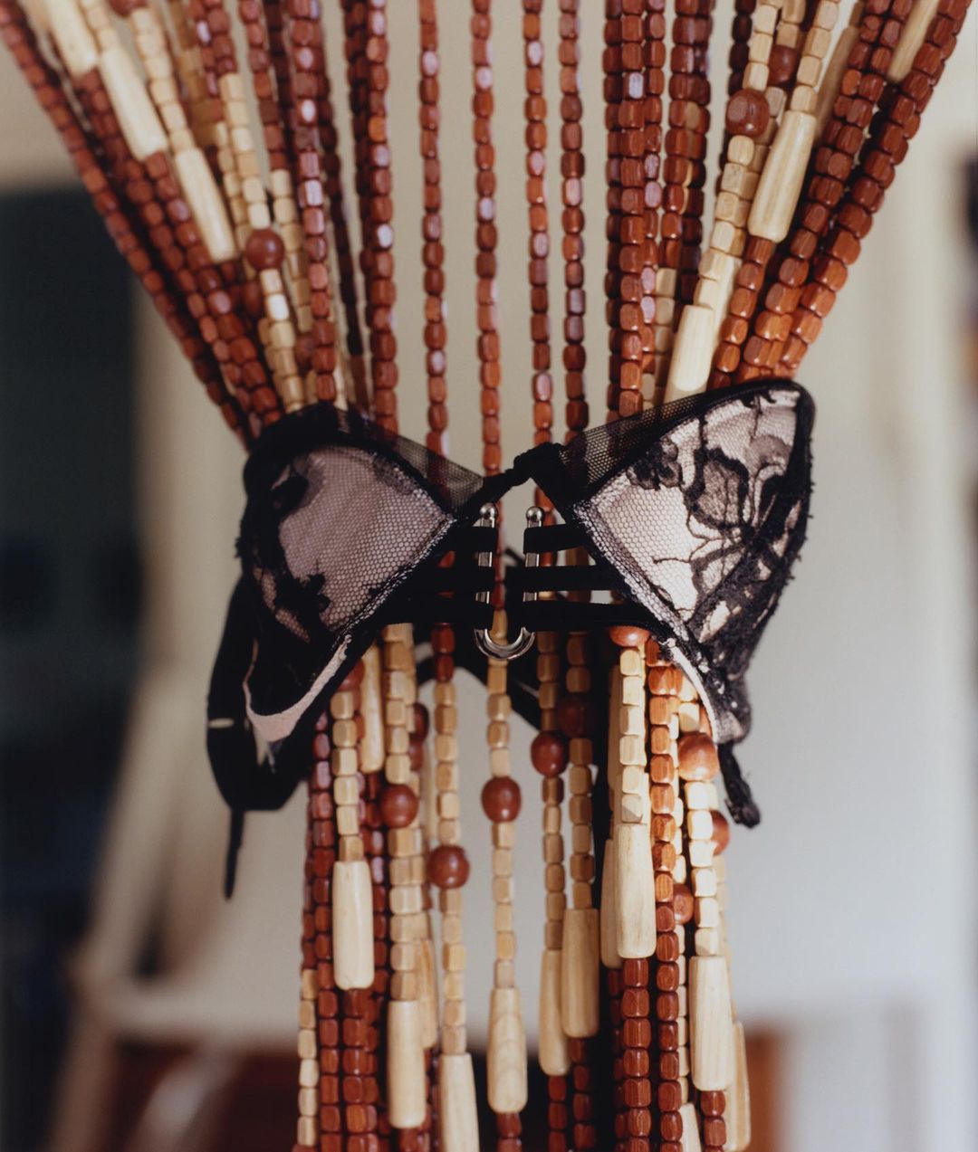 A close-up shot of a black lace bra tied to a beaded curtain with brown, tan, and white beads in an indoor setting with natural light.
