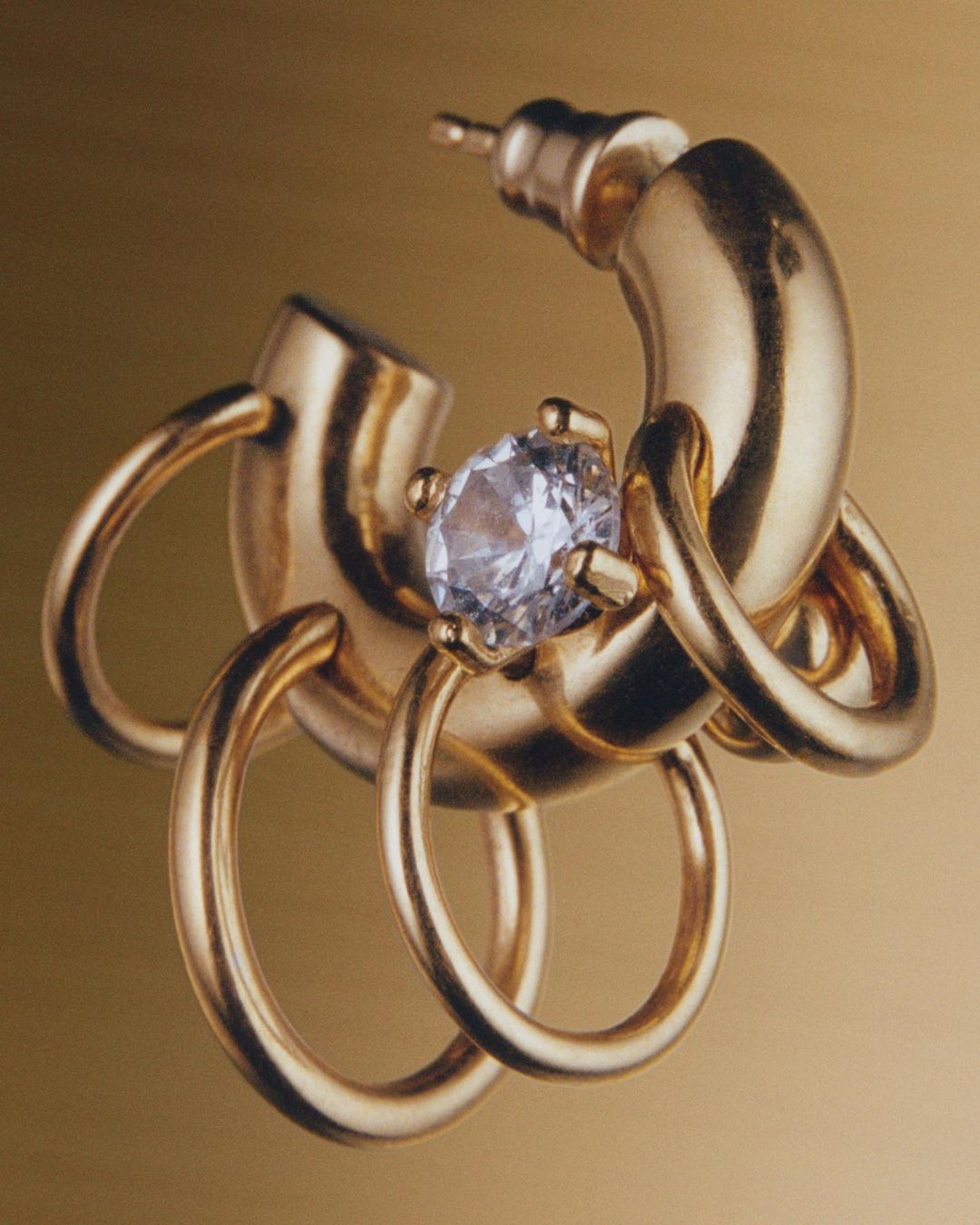 Close-up studio shot of a gold metal earring with a diamond set against a warm golden background. Fashion jewelry product shot.
