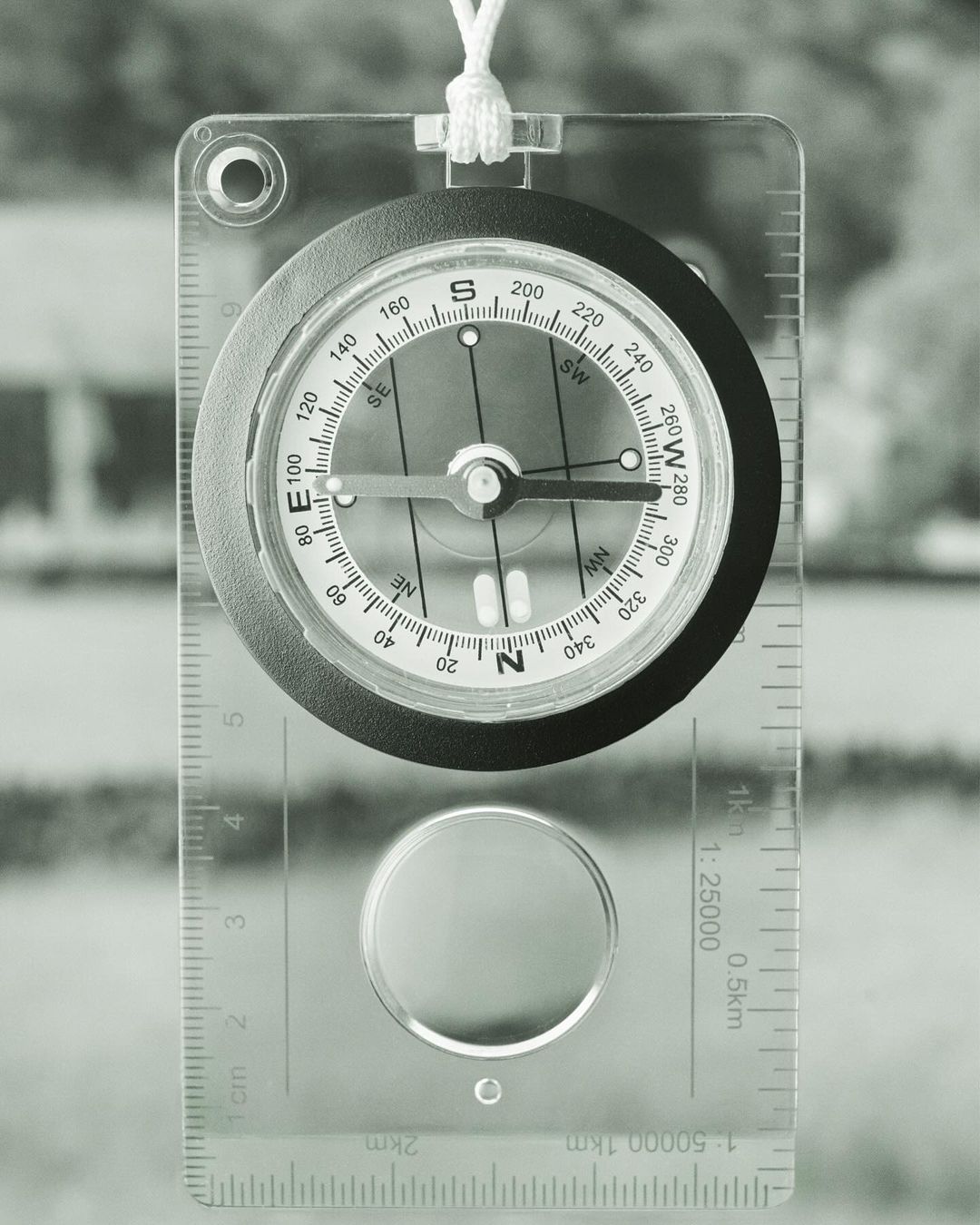 Overhead shot of a compass against a blurred background, showing direction, needle, and cardinal points with a clear casing and lanyard.
